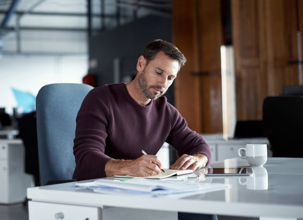 Man writing notes at desk with tablet and coffee