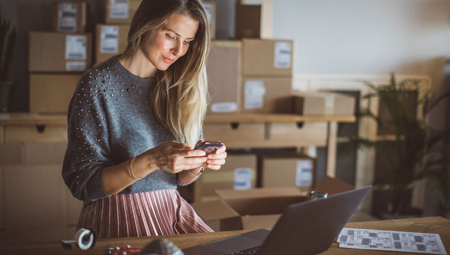 image for Woman looking at mobile banking app with laptop open on her desk